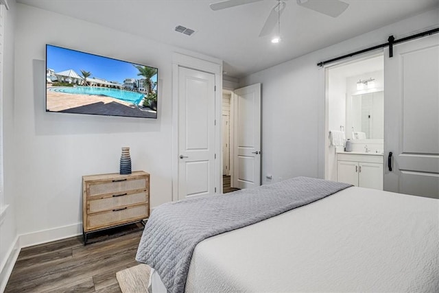 bedroom with ensuite bathroom, a barn door, dark wood-style flooring, a ceiling fan, and baseboards