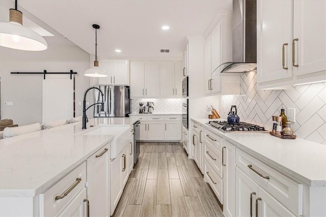 kitchen with tasteful backsplash, a barn door, appliances with stainless steel finishes, white cabinets, and wall chimney range hood