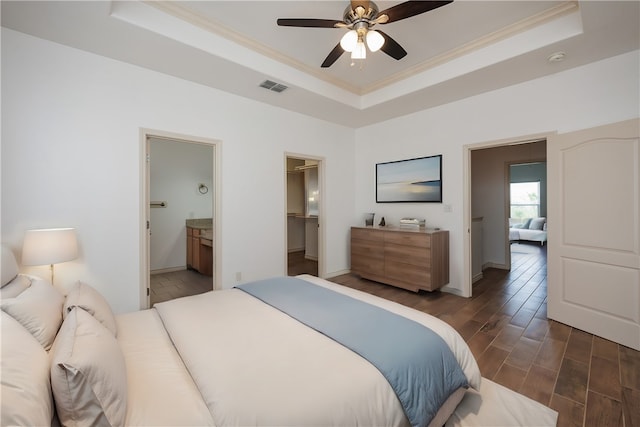 bedroom with crown molding, connected bathroom, dark wood-type flooring, a tray ceiling, and ceiling fan