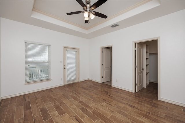 unfurnished bedroom with a spacious closet, crown molding, a raised ceiling, dark wood-type flooring, and ceiling fan