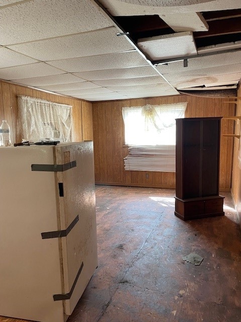 basement featuring white refrigerator, a drop ceiling, and wooden walls