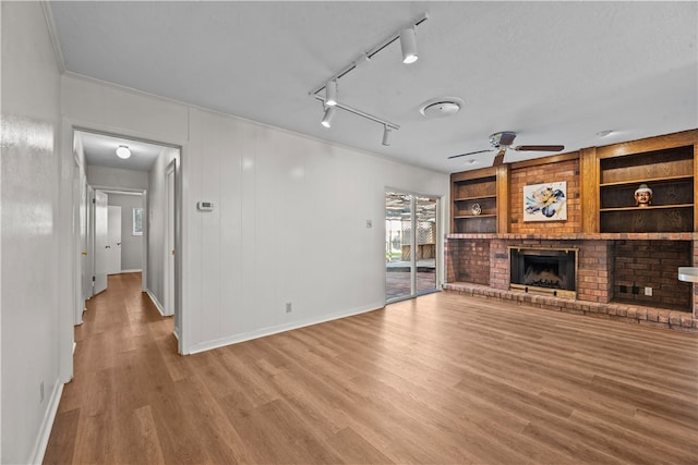 unfurnished living room with a brick fireplace, built in shelves, hardwood / wood-style floors, track lighting, and ceiling fan