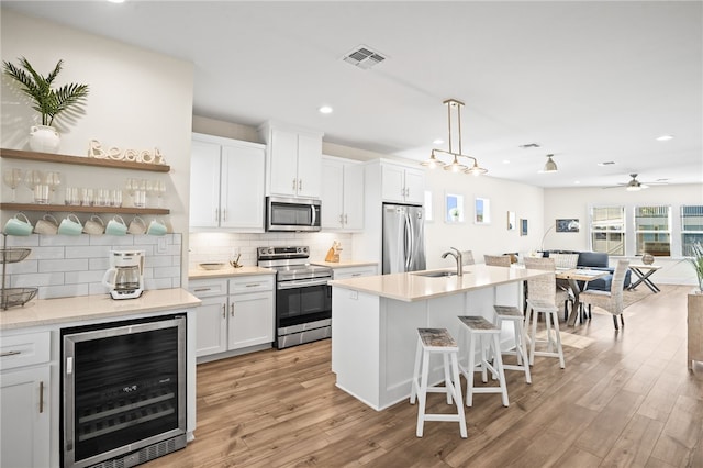 kitchen with appliances with stainless steel finishes, beverage cooler, a kitchen island with sink, light hardwood / wood-style floors, and white cabinetry