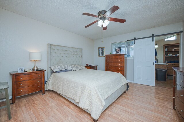 bedroom featuring connected bathroom, a barn door, a textured ceiling, light hardwood / wood-style floors, and ceiling fan