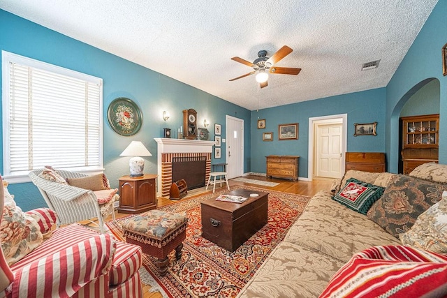 living room with a brick fireplace, visible vents, arched walkways, ceiling fan, and light wood-style floors