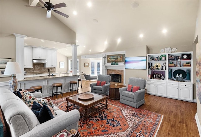 living area with a ceiling fan, wood finished floors, ornate columns, a fireplace, and high vaulted ceiling