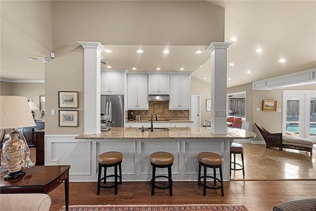 kitchen featuring stainless steel refrigerator, a kitchen breakfast bar, under cabinet range hood, ornate columns, and a sink