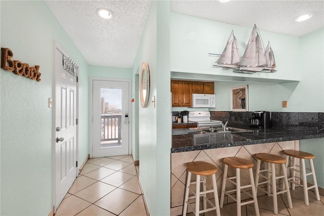 kitchen with light tile patterned floors, a peninsula, white appliances, a sink, and brown cabinets