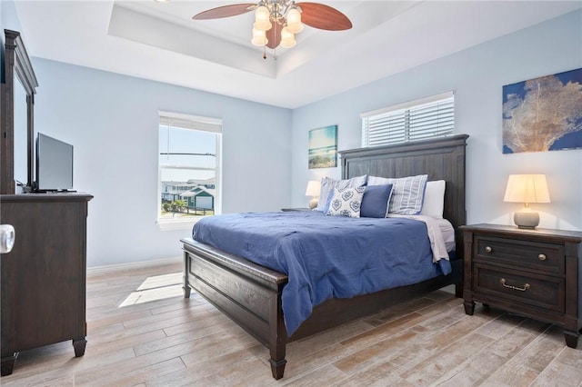 bedroom with a ceiling fan, light wood-style flooring, a tray ceiling, and baseboards