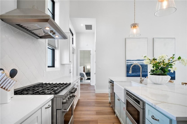 kitchen with decorative light fixtures, stainless steel appliances, a sink, light wood-type flooring, and wall chimney exhaust hood