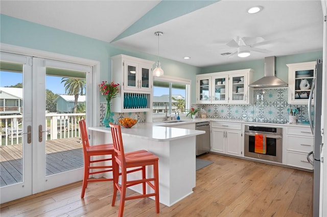 kitchen featuring white cabinets, decorative backsplash, a peninsula, stainless steel appliances, and wall chimney range hood