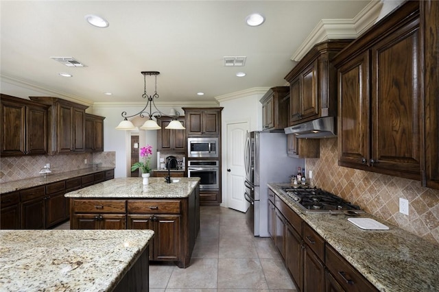 kitchen with appliances with stainless steel finishes, a kitchen island with sink, hanging light fixtures, dark brown cabinets, and light stone counters