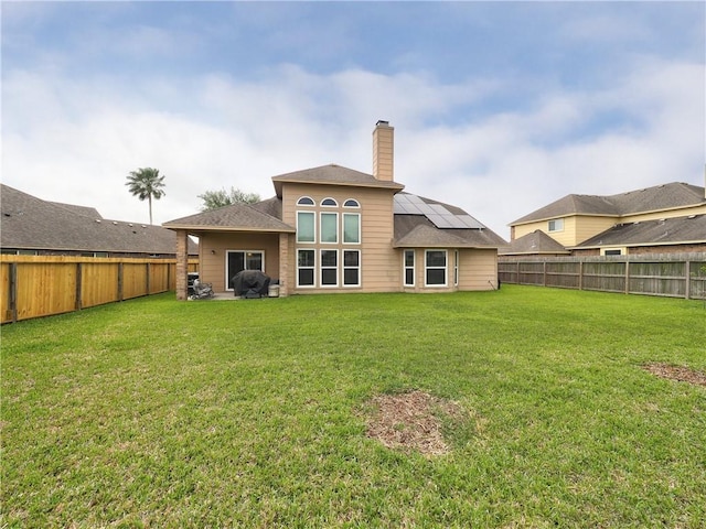 back of house with a fenced backyard, a chimney, a yard, and roof mounted solar panels