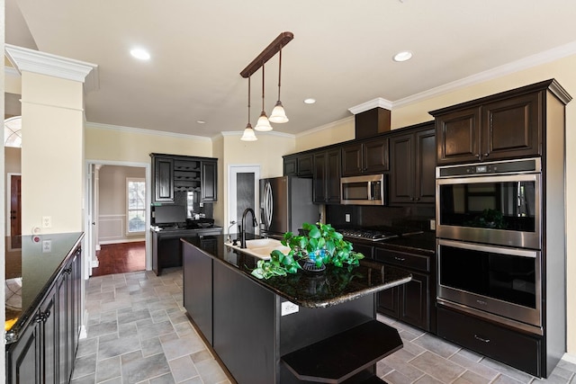 kitchen featuring sink, decorative columns, appliances with stainless steel finishes, dark stone counters, and a kitchen island with sink