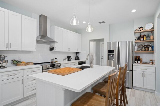 kitchen featuring a center island with sink, wall chimney range hood, tasteful backsplash, white cabinetry, and stainless steel appliances