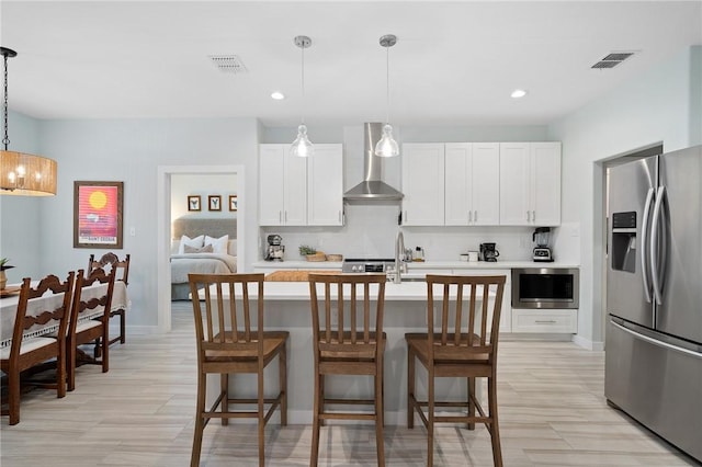 kitchen with a center island with sink, stainless steel fridge with ice dispenser, wall chimney exhaust hood, decorative light fixtures, and white cabinetry