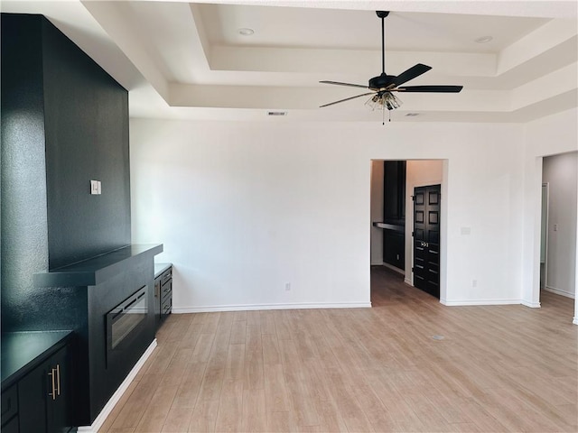 unfurnished living room with light wood finished floors, a tray ceiling, and baseboards