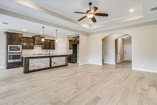 kitchen featuring an island with sink, backsplash, dark brown cabinetry, a tray ceiling, and stainless steel appliances