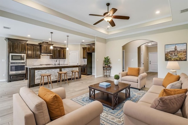 living room with sink, crown molding, light wood-type flooring, a raised ceiling, and ceiling fan