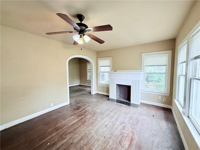 unfurnished living room featuring wood finished floors, a healthy amount of sunlight, arched walkways, baseboards, and a fireplace with flush hearth