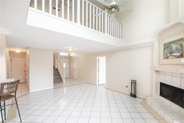 unfurnished living room with a high ceiling, stairway, a tiled fireplace, light tile patterned floors, and a ceiling fan