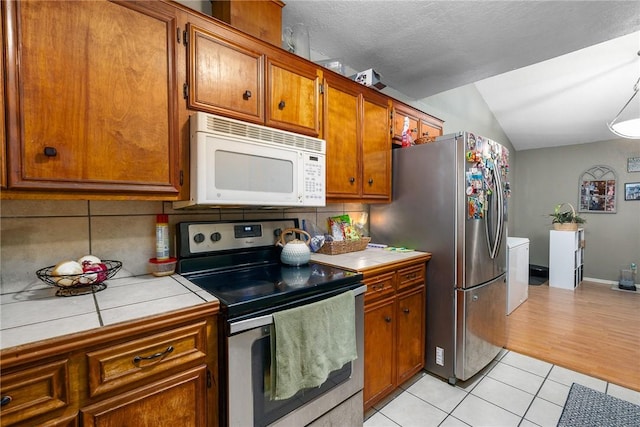 3713 Tripoli Dr, Corpus Christi, TX 78415 - kitchen with stainless steel appliances, tile counters, brown cabinets, vaulted ceiling, and light tile patterned flooring
