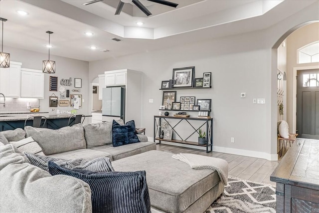 living room with ceiling fan, arched walkways, a tray ceiling, light wood-style flooring, and recessed lighting