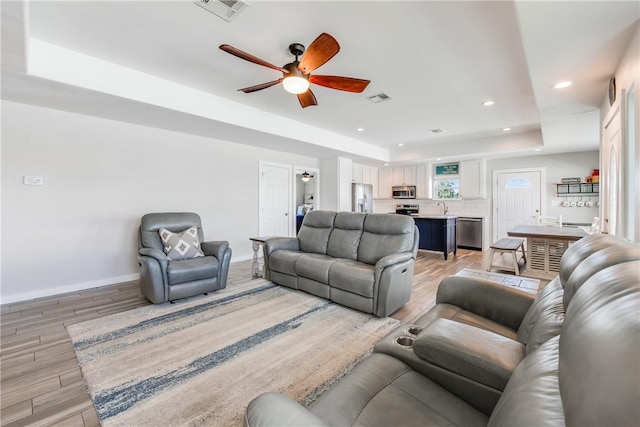 living room featuring ceiling fan, a tray ceiling, sink, and light wood-type flooring