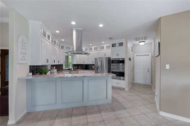 kitchen with island exhaust hood, appliances with stainless steel finishes, light stone countertops, white cabinets, and kitchen peninsula