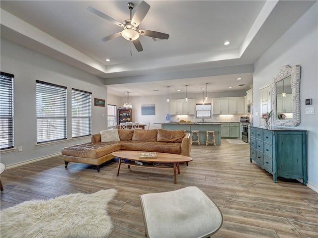 living room featuring a tray ceiling, a ceiling fan, light wood-style floors, baseboards, and recessed lighting
