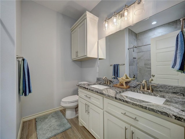 bathroom featuring wood finished floors, toilet, a tile shower, baseboards, and double vanity