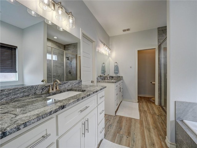 bathroom with wood finished floors, a shower stall, two vanities, and a relaxing tiled tub