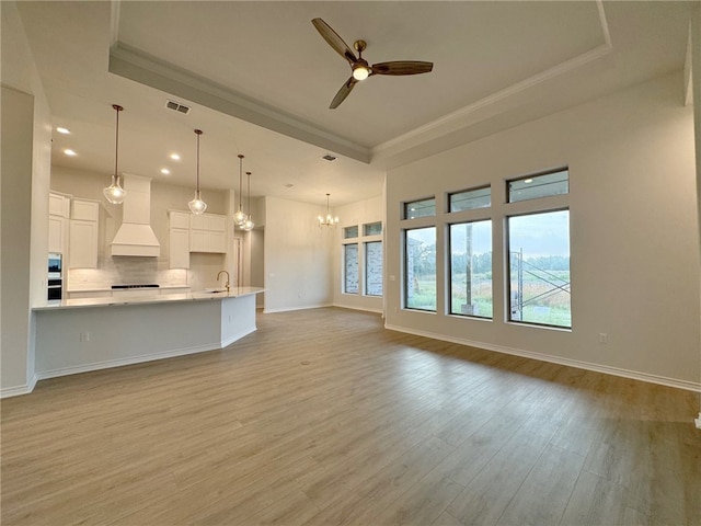 unfurnished living room with ceiling fan with notable chandelier, a tray ceiling, light hardwood / wood-style flooring, and crown molding