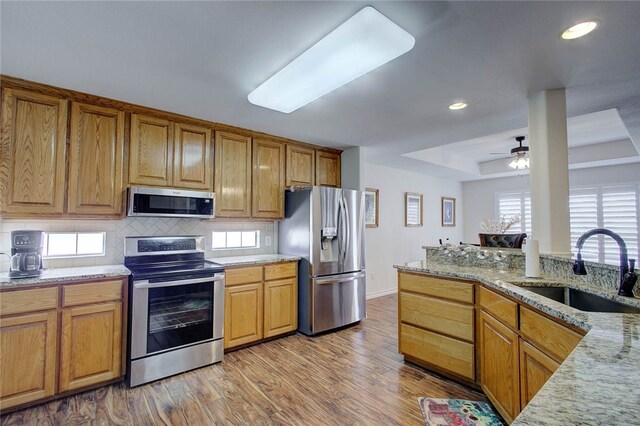 kitchen with stainless steel appliances, sink, a tray ceiling, light stone countertops, and light hardwood / wood-style flooring