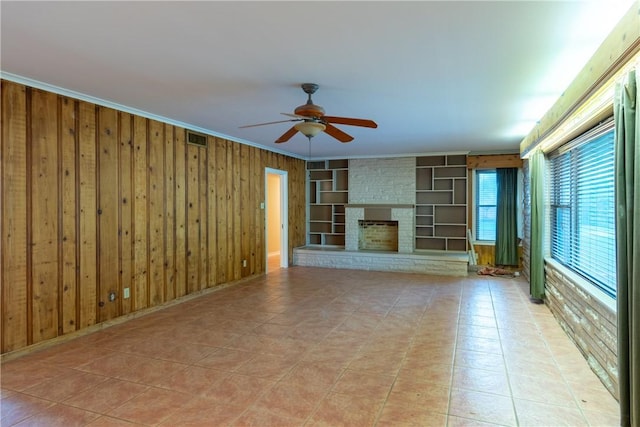 unfurnished living room with built in shelves, ceiling fan, wooden walls, a fireplace, and ornamental molding