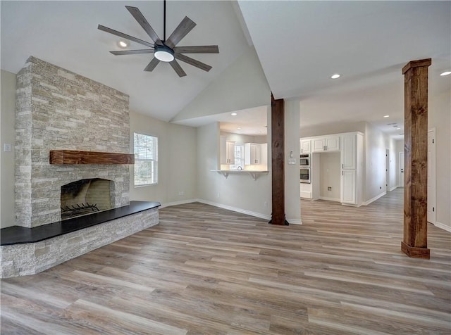 unfurnished living room featuring high vaulted ceiling, a stone fireplace, light hardwood / wood-style flooring, ceiling fan, and ornate columns