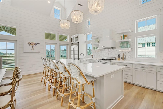 kitchen featuring built in fridge, decorative light fixtures, white cabinetry, wall chimney range hood, and a center island with sink