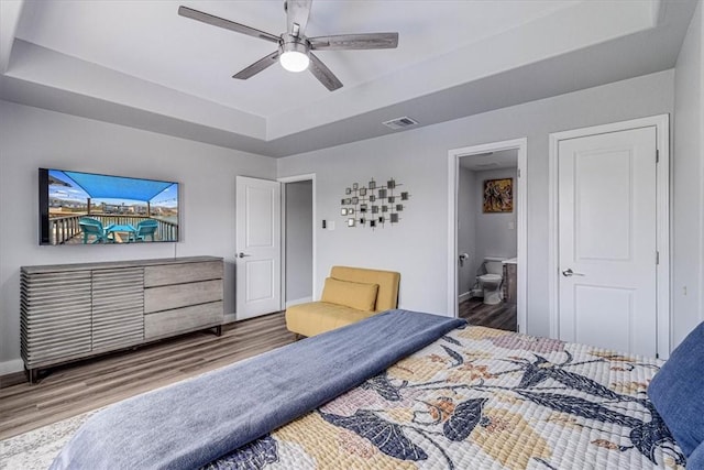 bedroom with wood finished floors, visible vents, baseboards, ensuite bath, and a tray ceiling