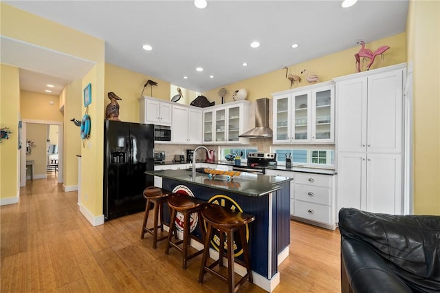 kitchen with a kitchen island with sink, black appliances, white cabinets, wall chimney exhaust hood, and a breakfast bar area