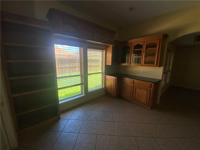 kitchen featuring light tile patterned floors, glass insert cabinets, brown cabinets, and dark countertops