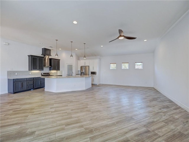 kitchen with wall chimney range hood, open floor plan, stainless steel appliances, a ceiling fan, and a center island with sink