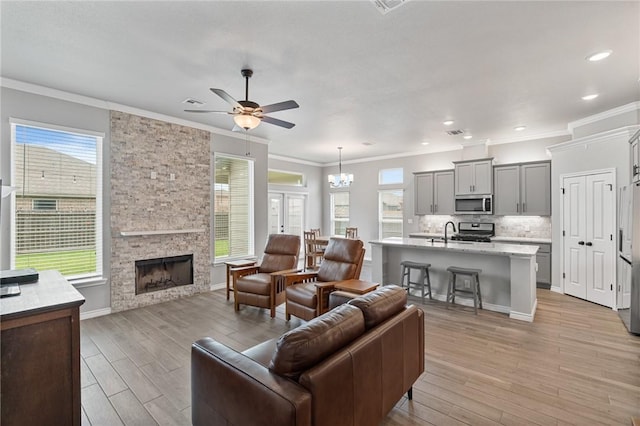 living room featuring crown molding, ceiling fan with notable chandelier, light wood finished floors, and a stone fireplace