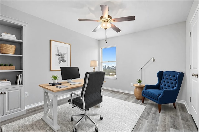 office space featuring ceiling fan, baseboards, built in shelves, and light wood-style flooring