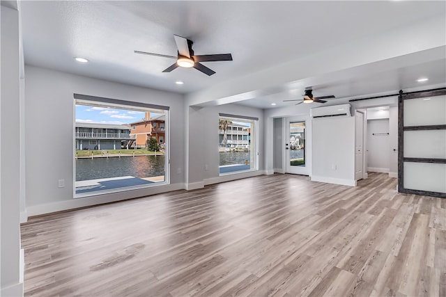 unfurnished living room featuring a water view, a barn door, ceiling fan, light wood-type flooring, and a wall mounted air conditioner