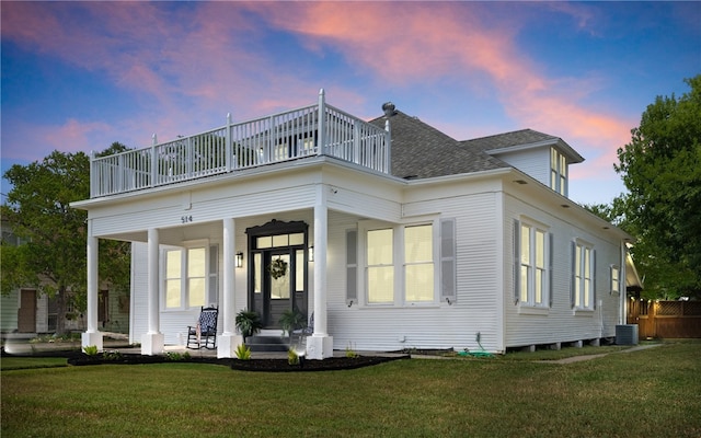 back house at dusk with a lawn, a porch, a balcony, and cooling unit