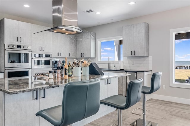 kitchen featuring a breakfast bar area, visible vents, stainless steel double oven, wall chimney range hood, and dark stone countertops