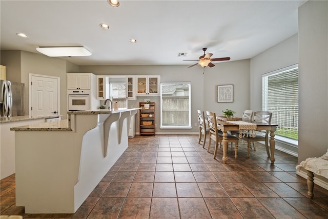 kitchen with light stone counters, white cabinets, oven, ceiling fan, and stainless steel refrigerator