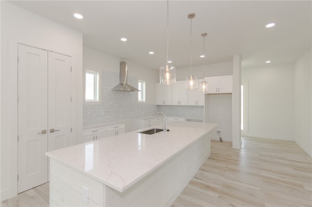 kitchen featuring a center island with sink, hanging light fixtures, sink, wall chimney range hood, and white cabinetry