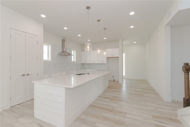 kitchen with light wood-type flooring, white cabinetry, pendant lighting, wall chimney exhaust hood, and a kitchen island with sink