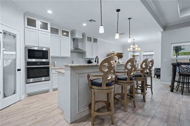 kitchen with light stone counters, an inviting chandelier, a kitchen island with sink, white cabinets, and wall chimney range hood